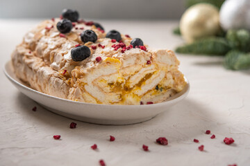 Homemade Meringue roll cake with cream, berries and almond on plate, selective focus, in the background Christmas tree branches with toys