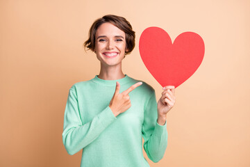 Photo of young attractive positive woman smile point finger paper heart advert promo recommend isolated over beige color background