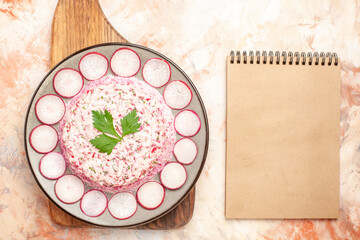 Horizontal view of delicious chicken salad with beet served with chopped red radishes on a gray plate on wooden cutting board and notebook on mixed color background