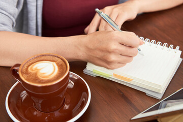 Close-up image of businesswoman drinking morning coffee in coffeeshop and writing thoughts and...