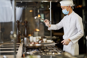 Skillful cook in face mask holding ladle over a pan