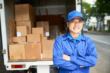Portrait of smiling young delivery truck driver in blue uniform standing with arms folded and...