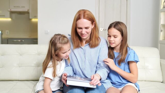 mom and daughters leaf through the photobook sitting at home on the couch.