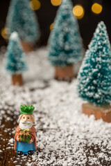 Close-up of figure of the Wise King Melchior with artificial Christmas trees on wooden table with snow, selective focus, with black background with lights in bokeh, vertical