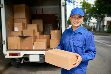 Portrait of cheerful Vietnamese delivery man with cardboard box standing at cargo van