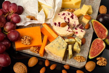 Cheese plate. Cheddar, Brie, Red Leicester, Wensleydale with sweetened dried cranberries, Blue Stilton, almond and walnut on a marble board with grape and figs on a black background.