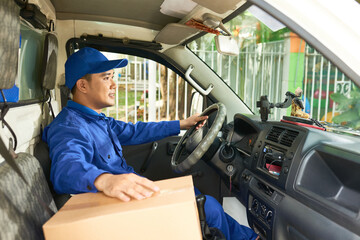Smiling positive courier in uniform driving delivery truck with cardboard box on front seat