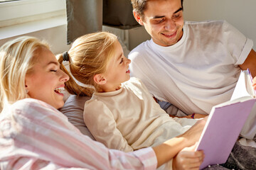 Obraz premium optimistic parents read book with child on bed before going to sleep, have talk, discuss the tale