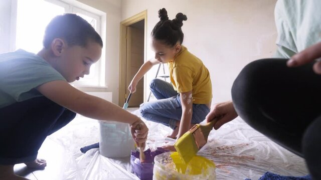 Cheerful teamwork family renovating flat. Funny kids move brushes in container with different color paint on floor with plastic cover at home during self isolation or holidays. Low angle shot 