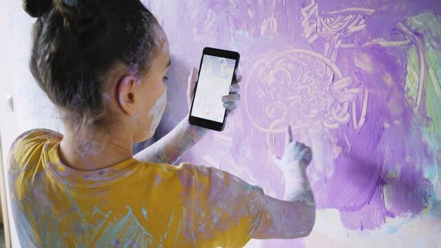 Children have fun in renovation room. Back view girl with paints on skin and clothes drawing picture your hands on wall and take video with black smartphone 