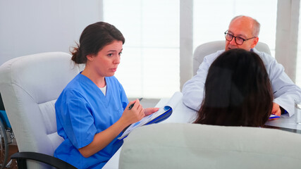 Obraz premium Young medical assistant briefing staff of doctors about patient diagnosis in conference room using clipboard. Clinic expert therapist talking with colleagues about disease, medicine professional