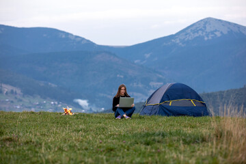 young female freelancer working on laptop in the mountains in the evening. Tourist girl sitting near campfire and tent. Copy space