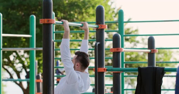 A man in a white shirt stretches his arms, pulls himself up on the bar, doing sports in office clothes on a street playground.
