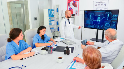 Obraz premium Mature medical physician explaining treatment to nurses during healthcare seminar pointing at digital monitor. Clinic herapist discussing with colleagues about disease, medicine professional.