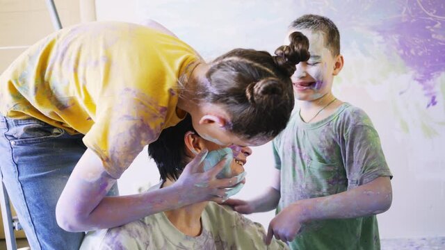 Boy and girl draw on mom face. Close-up funny children with paints on skin and clothes hug mother sitting on new apartment floor near window against coloured wall 