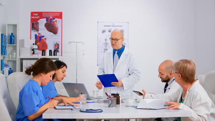 Senior specialist doctor presenting research informations to colleagues in hospital meeting room during brainstorming. Group of physicians talking about symptoms of disease in clinic office