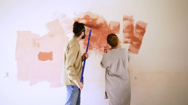 Couple have fun while renovating their apartment. young woman in grey shirt approaches bearded guy painting wall with roller lady checks palette kisses with laugh