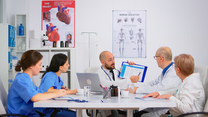 Doctor is brainstorming to analyze medical problems pointing on clipboard showing to colleagues medical statistics. Coworkers working in hospital office discussing symptoms of disease, taking notes.