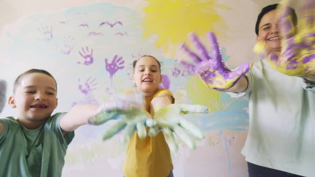 funny children with mother in white t-shirt show smeared hands with various paints against coloured wall in new apartment, family celebrating moving day close view