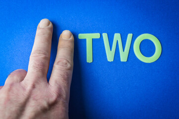 Two human finger beside the word Two written with plastic letters on blue paper background, concept
