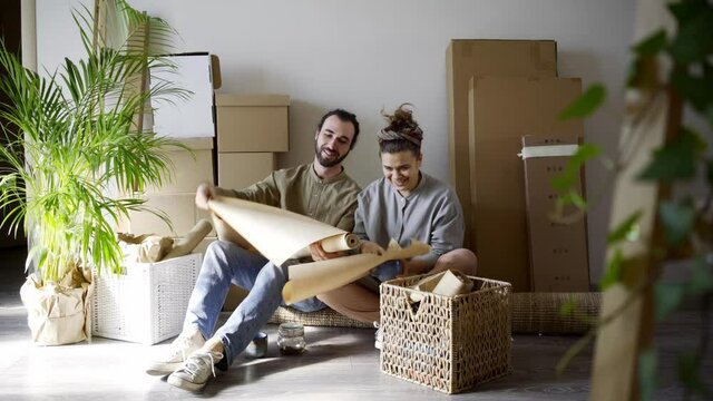 joyful family of happy husband and wife wearing casual clothes tear up craft paper for packing different belongings and put into box while sitting on floor to remove to new flat