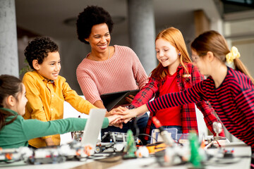 Happy kids with their African American female science teacher with laptop programming electric toys and robots at robotics classroom