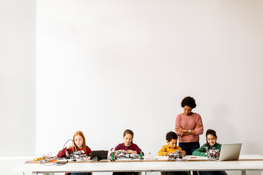 Happy Kids With Their African American Female Science Teacher With Laptop Programming Electric Toys And Robots At Robotics Classroom