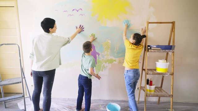 joyful family painting wall drawing images with hands and colorful paints at new room near window at bright day light for celebrating children party back view