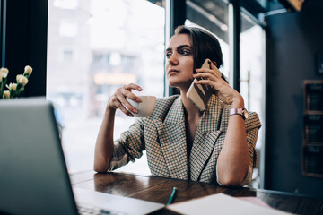 Smart casual manager with coffee cup making business conversation via smartphone application for discussing delivery order, Caucasian female small owner sitting at cafe table with laptop and calling