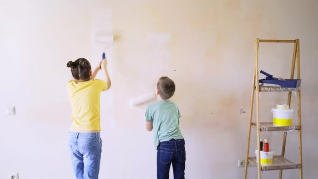  joyful girl and boy painting new apartment wall with rollers, happiness standing near wooden ladder with tools medium shot backside view. Finishing housework renovate concept
