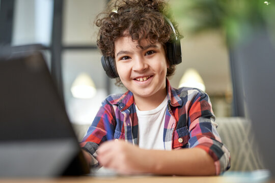 Cheerful latin american school boy wearing headphones, smiling at camera and making notes. Child using digital tablet while studying at home