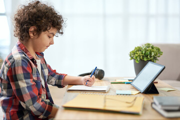 Enthusiastic latin school boy making notes, having online lesson using digital tablet while studying at home