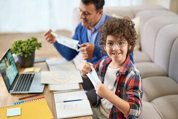 Keep safety in mind. Cheerful latin boy smiling at camera, wearing protective mask while sitting at the desk together with his father, using laptop, having online lesson indoors