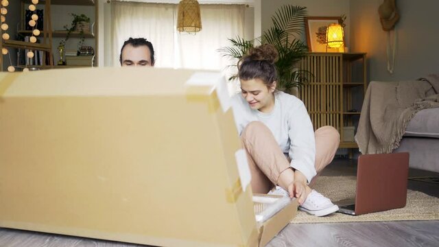Millennial young couple unpacking new furniture. Bearded man and cheerful woman take out parts of new table from big cardboard box while sitting on floor in spacious light living room