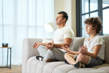 Peaceful vibes. Relaxed latin middle aged father and his son meditating together on a couch at home