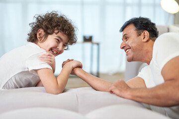 Competition. Cute latin boy and his father having fun together at home while armwrestling lying on the couch