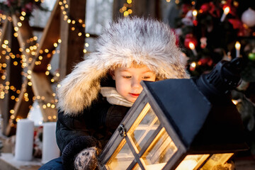 A little boy in winter clothes holds a Christmas lantern in his hands and looks at it. Outdoors, in winter.