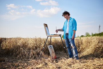 Full-length portrait of young male artist wearing blue shirt, drawing on canvas on easel on wheat...