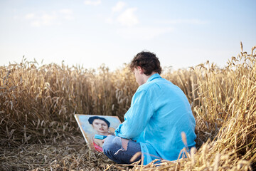 Naklejka na ściany i meble Young male artist, wearing blue shirt and torn jeans, sitting on wheat field, drawing on canvas. Painting workshop in rural countryside. Artistic education concept. Outdoors leisure activities.