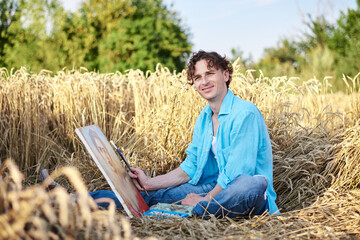 Naklejka na ściany i meble Young male artist, wearing blue shirt and torn jeans, sitting on wheat field, drawing on canvas. Painting workshop in rural countryside. Artistic education concept. Outdoors leisure activities.