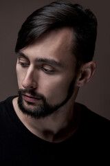 Portrait of handsome caucasian man with beard and stylish haircut in black clothes posing on brown background isolated in studio, fashion model lowering eyes