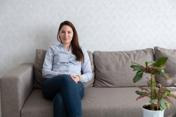 A young beautiful woman in a blue shirt and jeans is sitting on the couch and looking at the camera. Girl sitting on the sofa