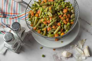 Overhead shot of steamebeans and carrots in an iron strainer on a kitchen table