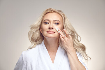 Good looking mature woman in spa bathrobe applies cream to the face over beige isolated.