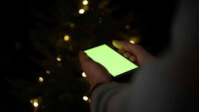 Holding Smartphone with greenscreen in front of christmas tree. Iphone 12 with chroma key background for inserting custom content like gifts, website, application.