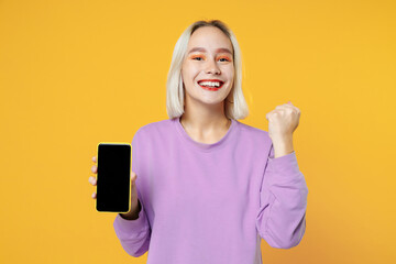 Young excited caucasian woman in basic purple shirt hold mobile phone blank screen workspace copy space mock up showing winner gesture clenching fist isolated on yellow background studio portrait.