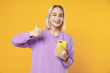 Young blonde caucasian gladden woman 20s bob haircut wearing casual basic purple shirt holding mobile cell phone gadget showing thumbs up gesture isolated on yellow color background studio portrait.