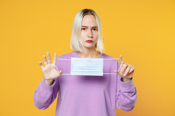 Young serious caucasian woman 20s in basic purple shirt holding sterile face mask to safe from coronavirus virus covid-19 during pandemic quarantine isolated on yellow background studio portrait.