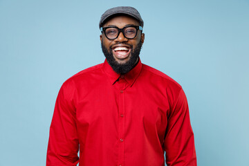 Cheerful laughing handsome young bearded african american man 20s wearing casual red shirt cap eyeglasses standing and looking camera isolated on pastel blue color wall background studio portrait.