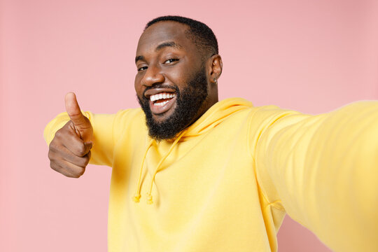 Close up of cheerful young african american man 20s wearing casual basic yellow streetwear hoodie doing selfie shot on mobile phone showing thumb up isolated on pastel pink background studio portrait.